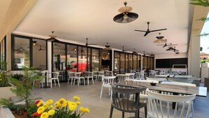 An outdoor dining area with multiple tables and chairs, ceiling fans, and a garden with yellow flowers. - Hotel Tina (Corfu)