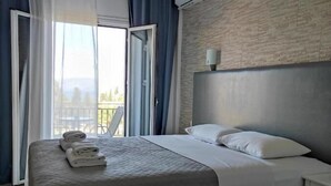 A hotel room with a bed, a nightstand, a wall-mounted air conditioner, and a view of the outdoors through a large window with curtains. - Hotel Tina (Corfu)