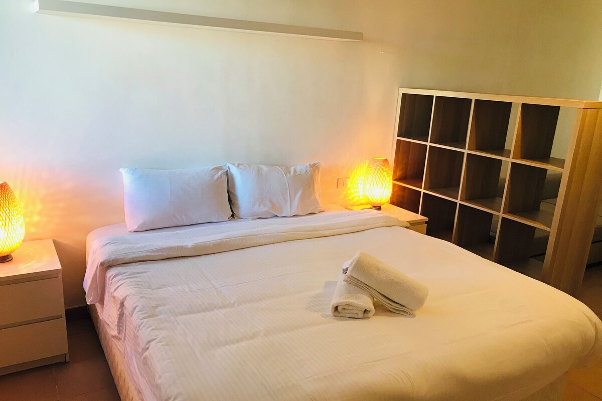 Una cama bien hecha con almohadas y una toalla doblada, dos lámparas de noche y un armario de madera con varios compartimentos.