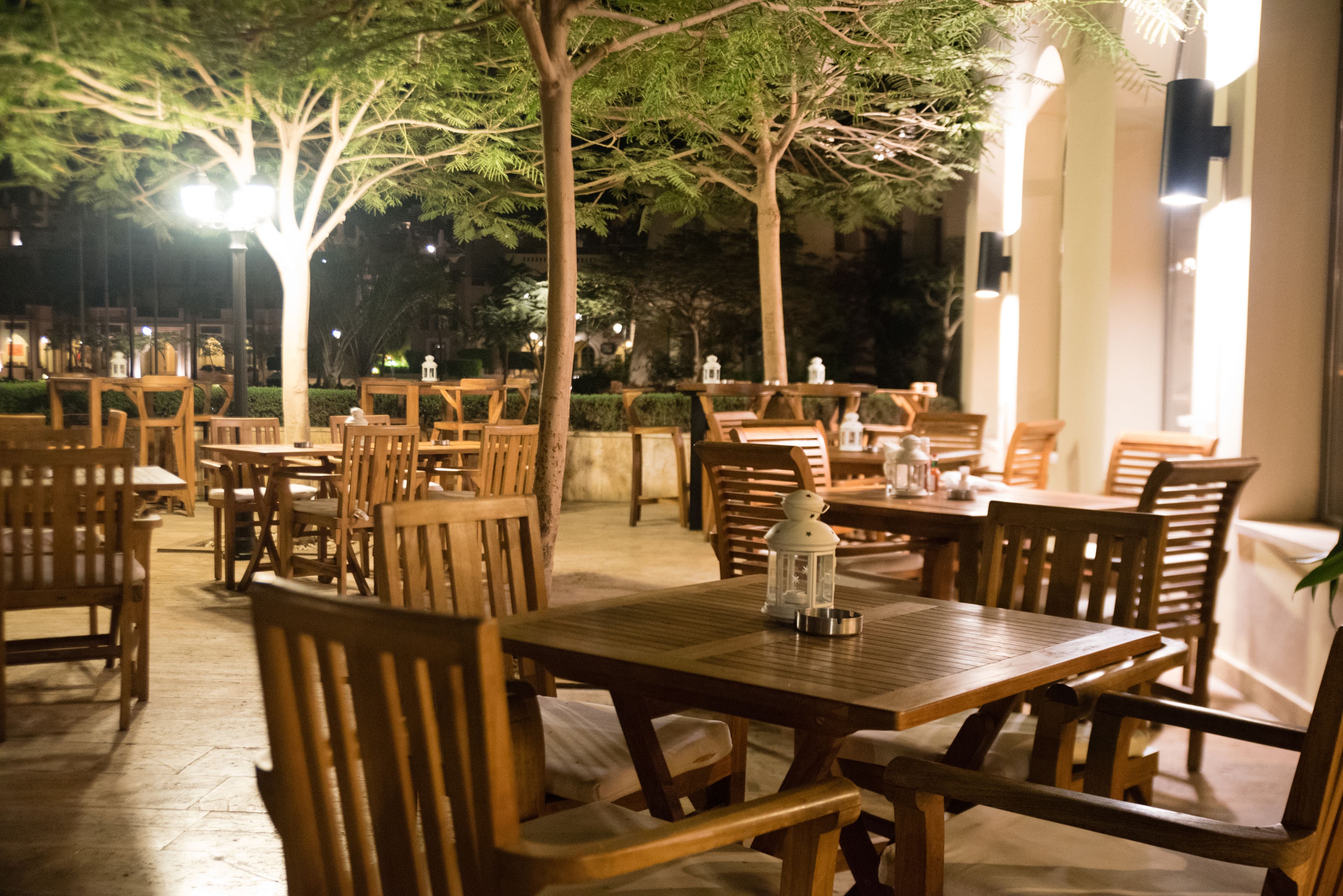 An outdoor dining area with wooden tables and chairs, surrounded by trees and illuminated by soft lighting.