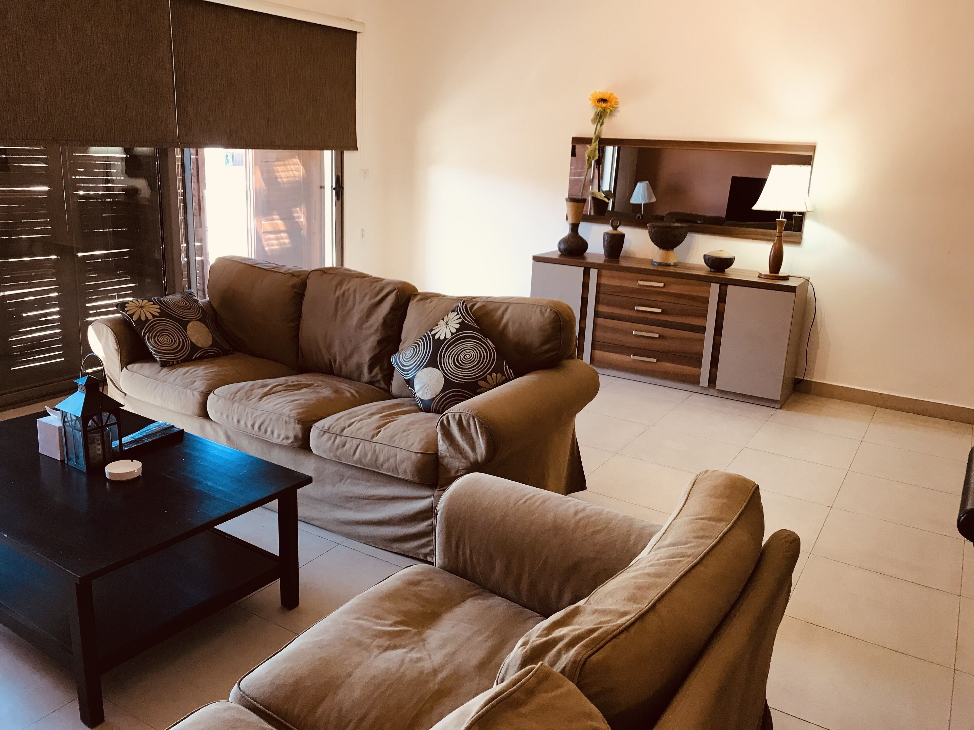 A living room with a sofa, a coffee table, and a side table. There is a lamp on a cabinet and a window with blinds.