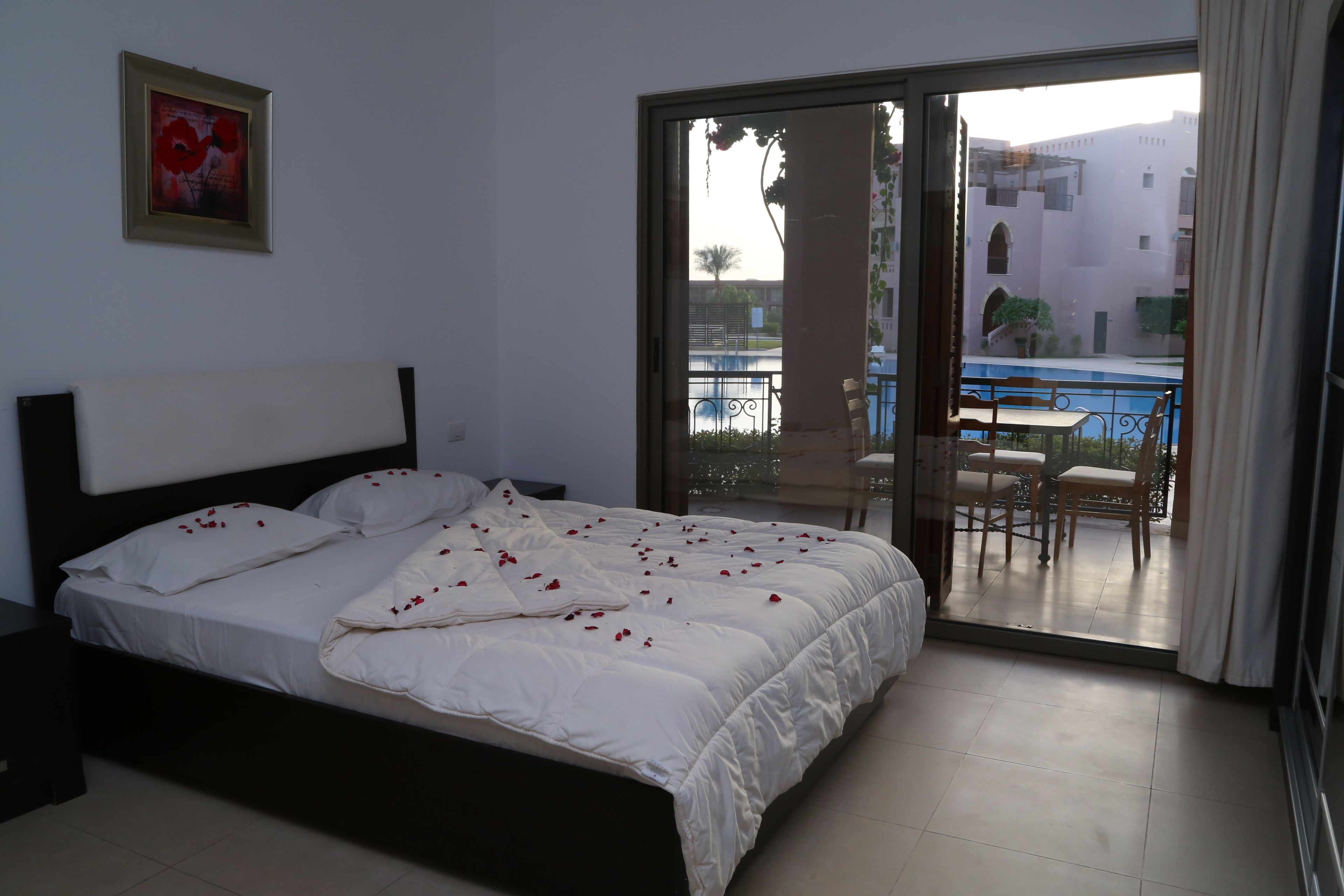 A hotel room with a bed, a painting on the wall, and a view of a pool and outdoor seating through a glass door.