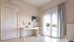 Ett hotellrum med en vit dörr, en platt-TV som är monterad på väggen, ett litet bord med en vas och glas, och ett stort fönster med gardiner.