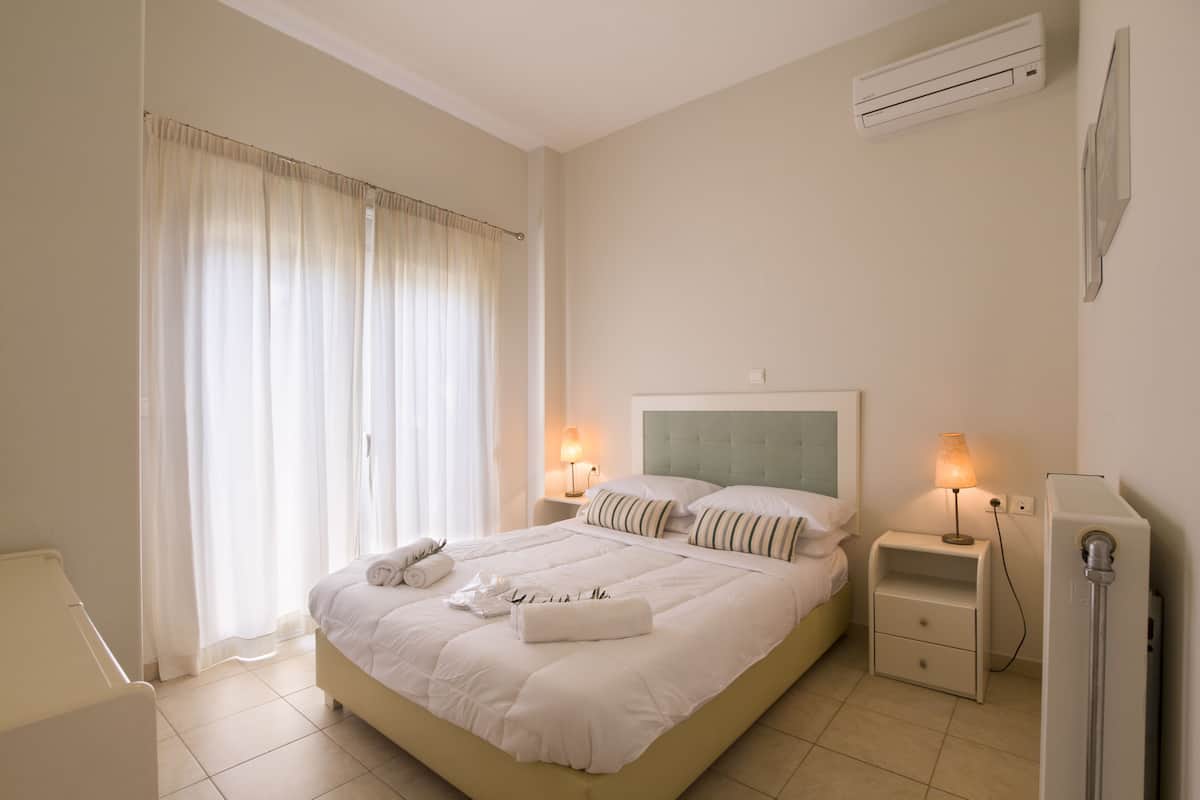 A neatly arranged hotel room with a large bed, two bedside tables with lamps, a window with curtains, and an air conditioning unit on the wall.