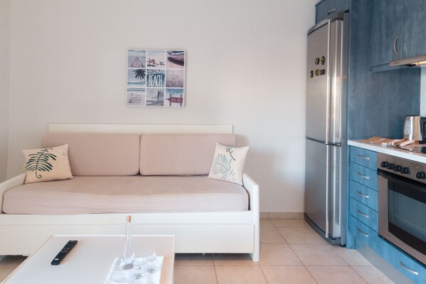 A compact living space with a sofa, a small table, a refrigerator, and a kitchen area. - Like Home (Platanias)