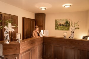 Uma mesa de recepção com painéis de madeira, uma pintura emoldurada de paisagem na parede e um vaso com flores brancas.
