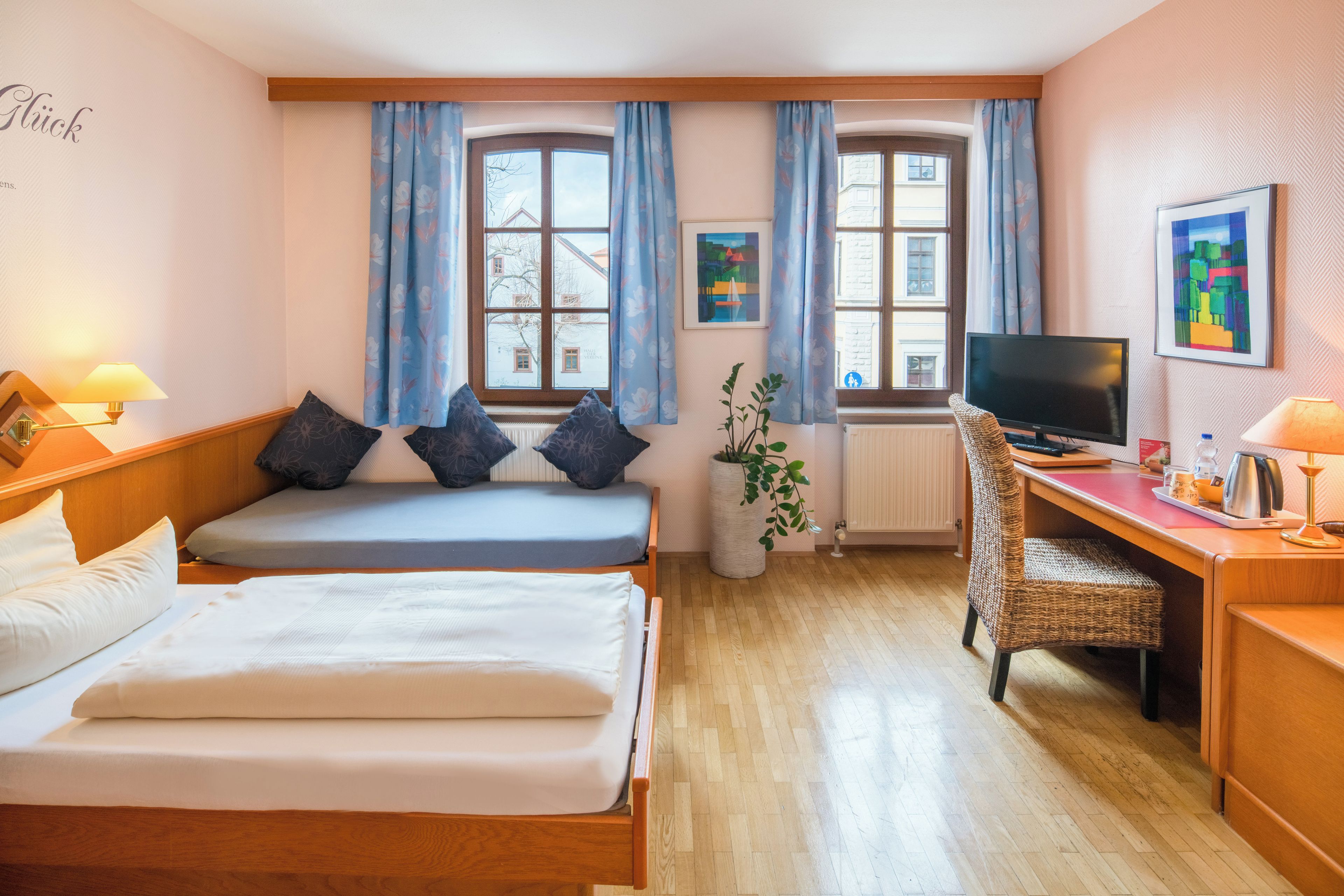A hotel room with two beds, a desk, a television, and large windows with curtains.