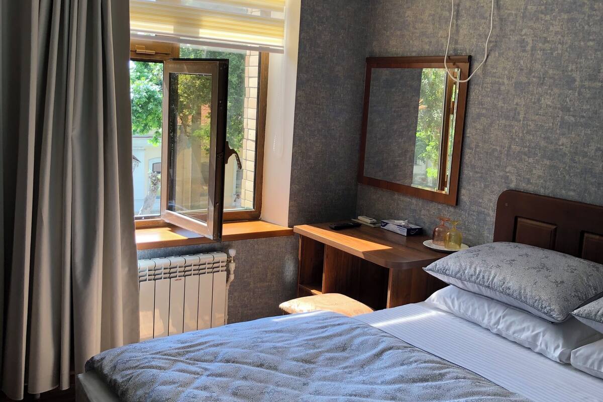 A hotel room with a bed, a desk, a radiator, an air conditioning unit, and a window with curtains.