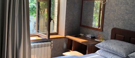 A hotel room with a bed, a desk, a radiator, an air conditioning unit, and a window with curtains.
