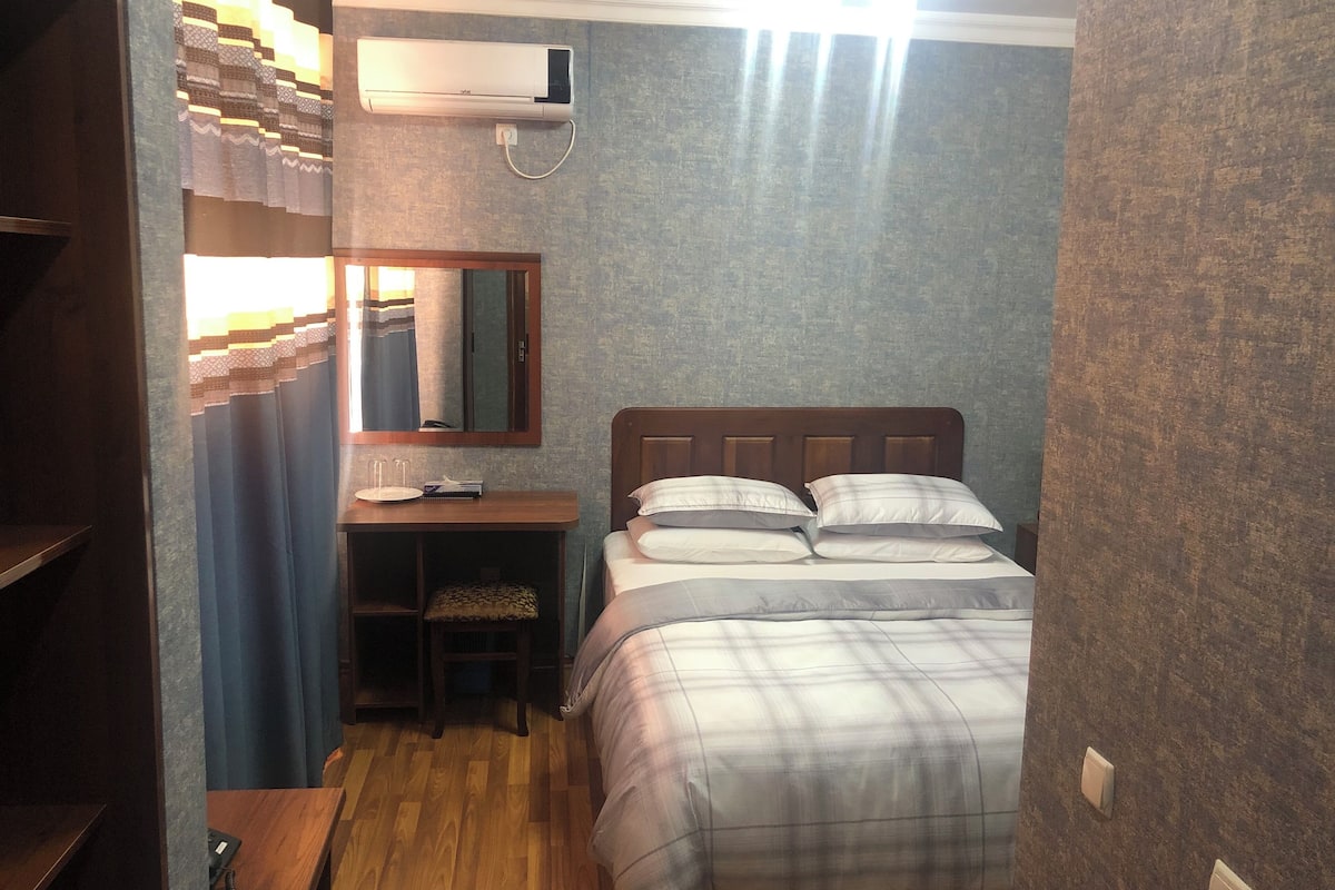 A compact hotel room with a bed, a bedside table, a desk, and an air conditioning unit.