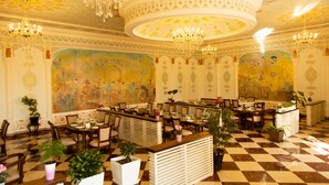 A luxurious hotel lobby with ornate chandeliers, checkered flooring, and detailed murals on the walls.