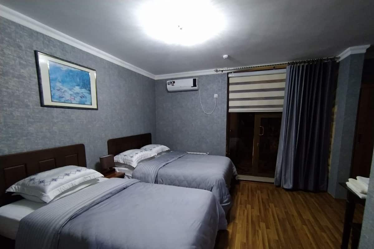 A hotel room with two beds, a ceiling light, an air conditioning unit, a window with curtains, and a wooden floor.