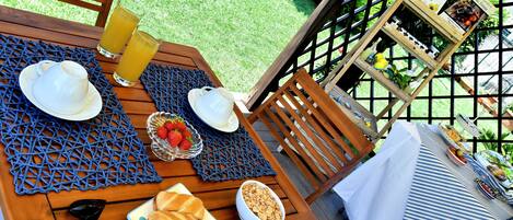 Una mesa de madera con cosas para el desayuno, como cruasanes, un tazón de cereal y vasos de jugo, en un patio con vista a un jardín.