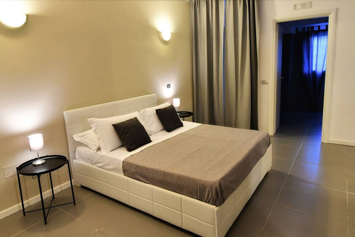 A neatly made bed with a white headboard and two black pillows, a brown bedspread, and two bedside tables with lamps.