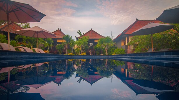 A pool with umbrellas and palm trees, surrounded by buildings with red-tiled roofs. - Perdana Lembongan (Lembongan Island)