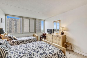 A bedroom with two beds, a dresser, a chair, a television, and a window with a city view.