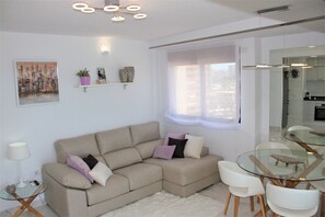 A modern living room with a beige sectional sofa, a glass coffee table, a dining area with a glass table and white chairs, a cityscape painting, and a small potted plant.