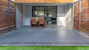 Uma área de pátio com duas cadeiras de madeira e uma mesa, uma porta de vidro que dá para o interior e piso ladrilhado.