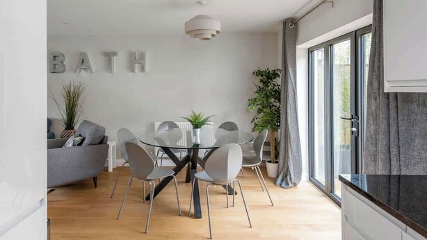 Un espace repas moderne avec une table et des chaises en verre, un canapé gris et un panneau indiquant « SALLE DE BAIN » sur le mur.