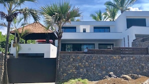 Une maison moderne à deux étages, dotée d’un mur en pierre et bordée de palmiers.