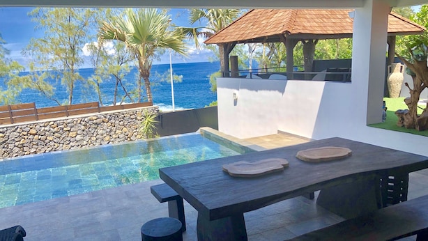 Une piscine à débordement offrant une vue sur l’océan, une table en bois et un mur en pierre.