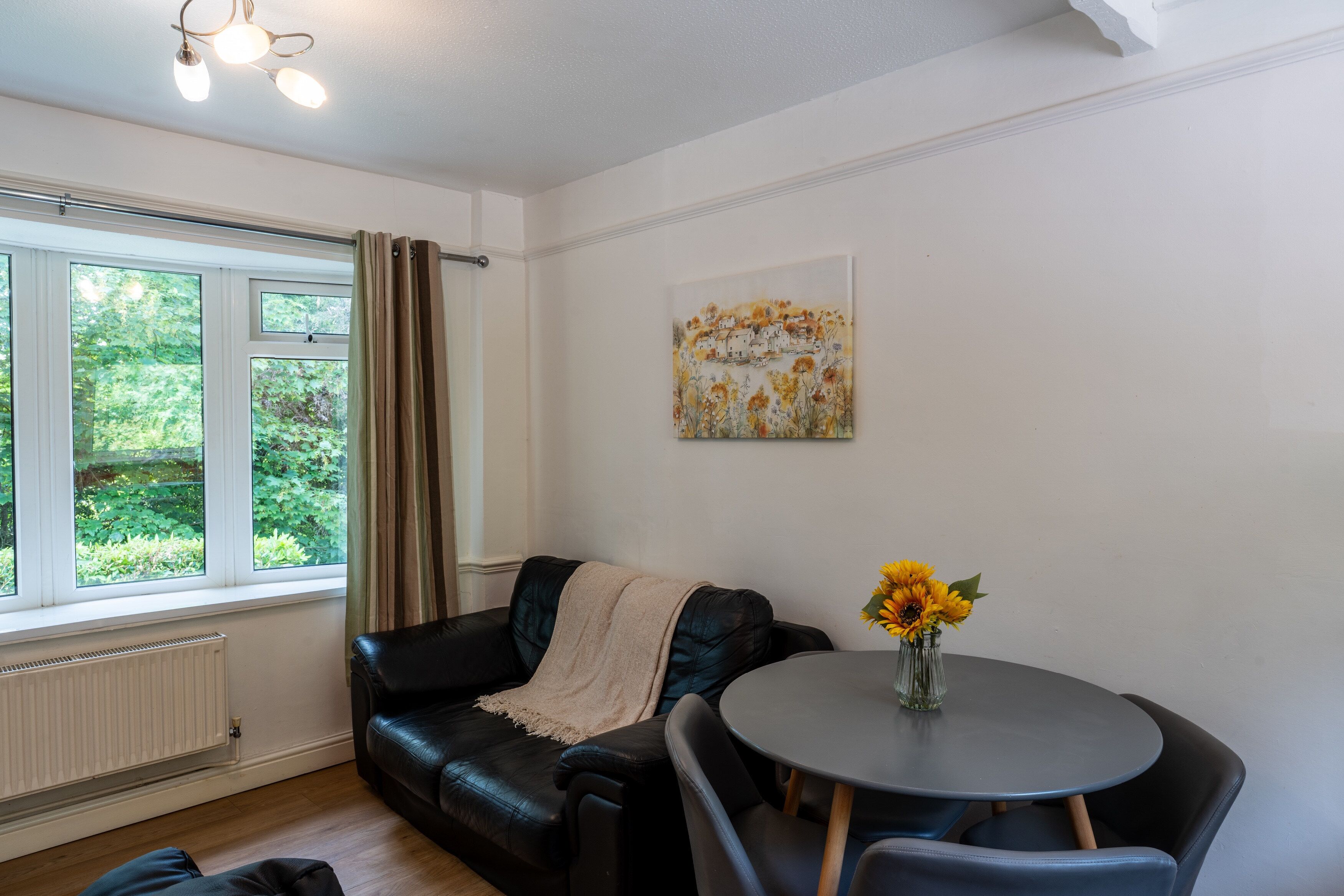 Una habitación con un sofá de cuero negro, una mesa redonda con un jarrón de girasoles, un radiador y una ventana con cortinas.