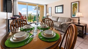 Un comedor con una mesa redonda preparada para cuatro personas, sillas, un sofá, un televisor y un balcón con vistas a palmeras.