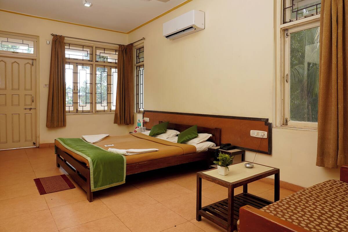 A bedroom with a bed, a bedside table, a window with curtains, an air conditioning unit, and a door.