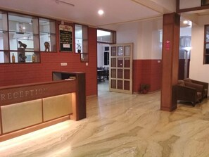 Un área de recepción con piso de mármol, un mostrador de recepción con la etiqueta “RECEPTION” y una vitrina de vidrio con objetos.