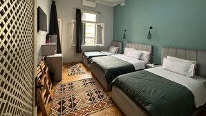 A bedroom with two beds, a desk, a chair, a window with curtains, a patterned rug, and a wall-mounted TV. - Cheers Hostel Cairo (Cairo)