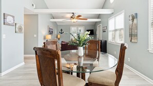 A dining area with a glass table surrounded by wooden chairs, a ceiling fan, and wall-mounted artwork.