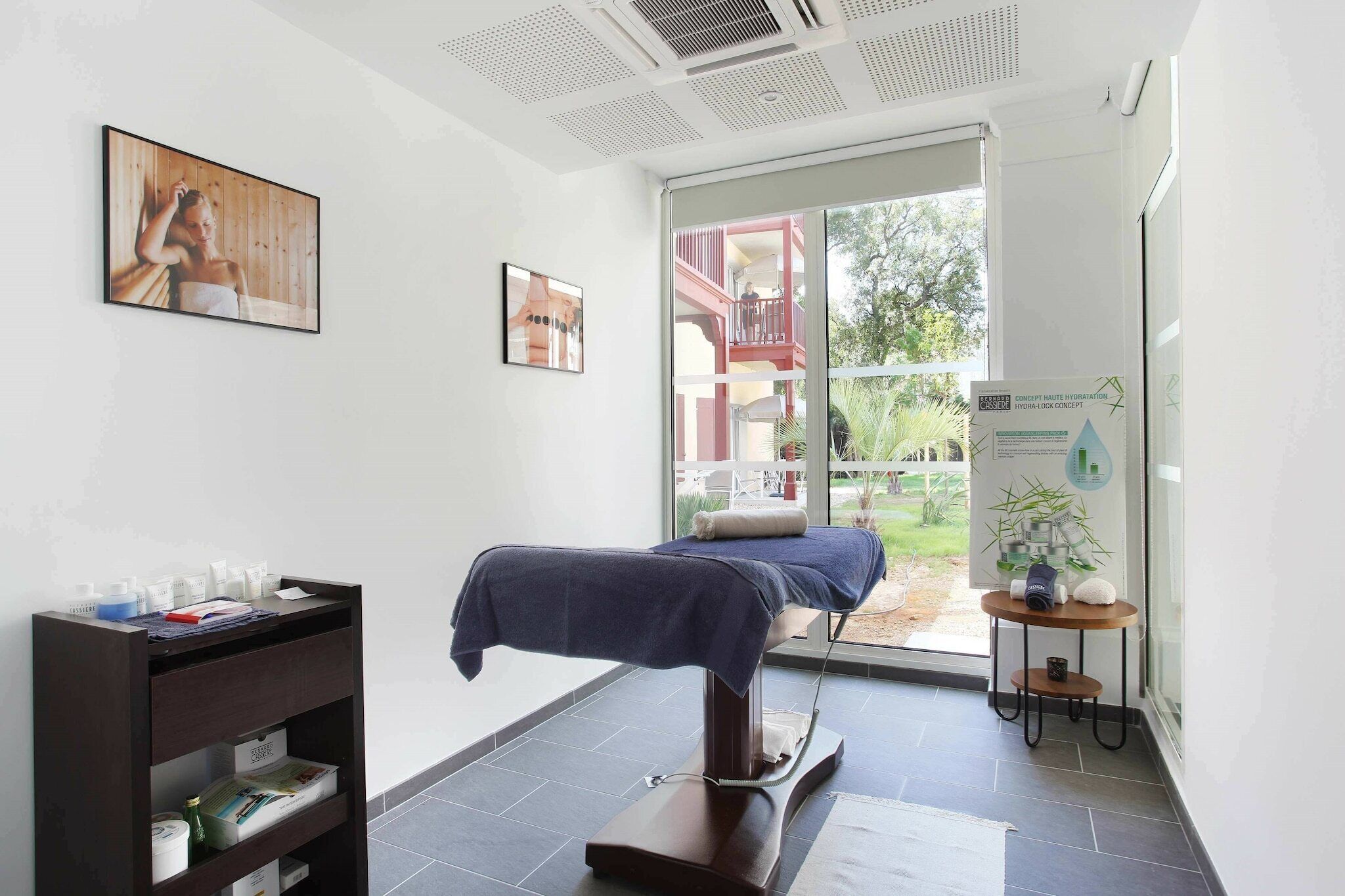 A room with a bed, a shelf with toiletries, a poster on the wall, and a window with a view of the outside.