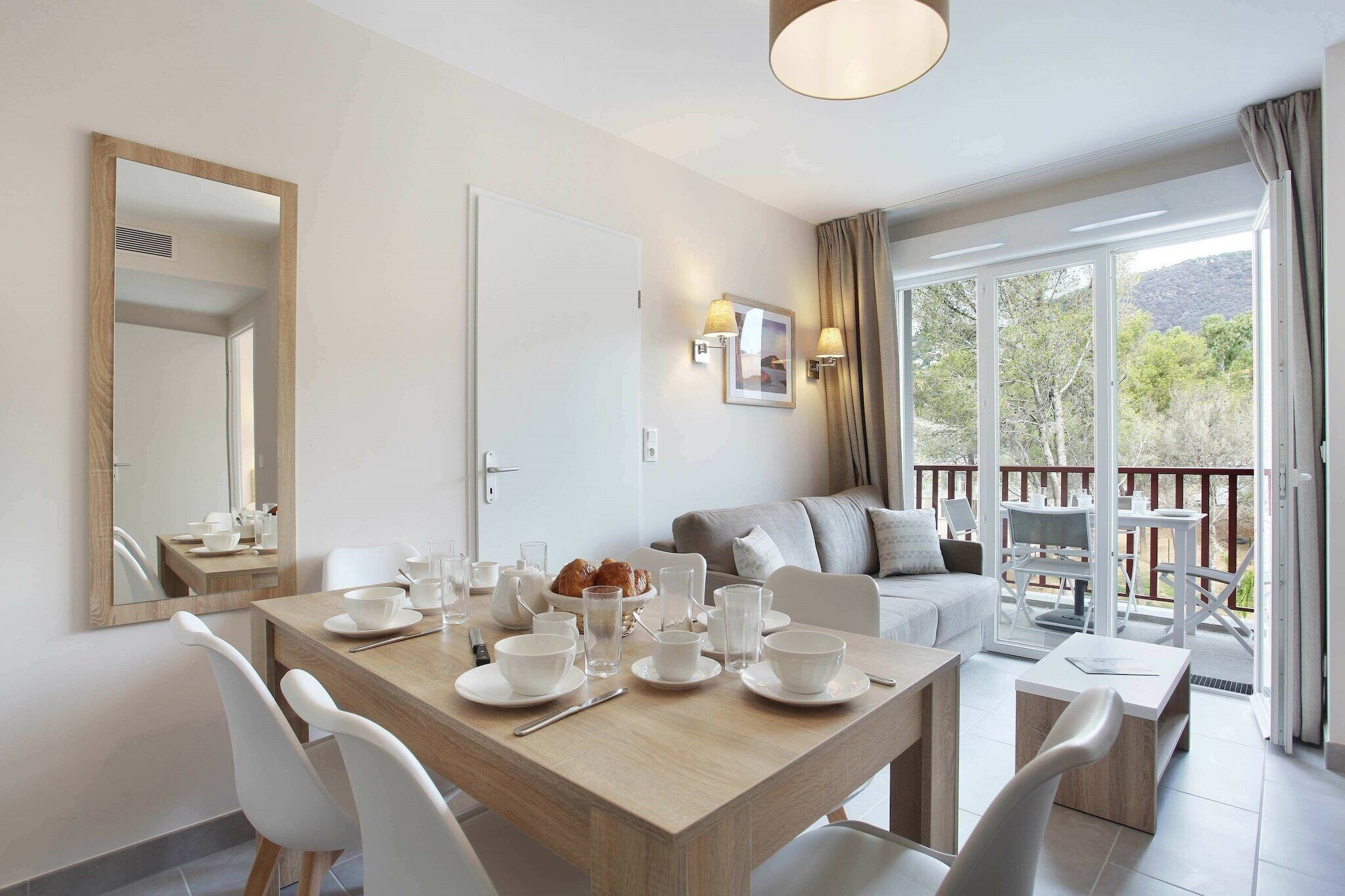 A modern dining area with a wooden table set for breakfast, a sofa, and a balcony with a view of trees and hills.
