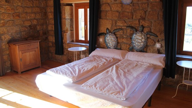A bedroom with a wooden bed, a small wooden dresser, and a stone wall.