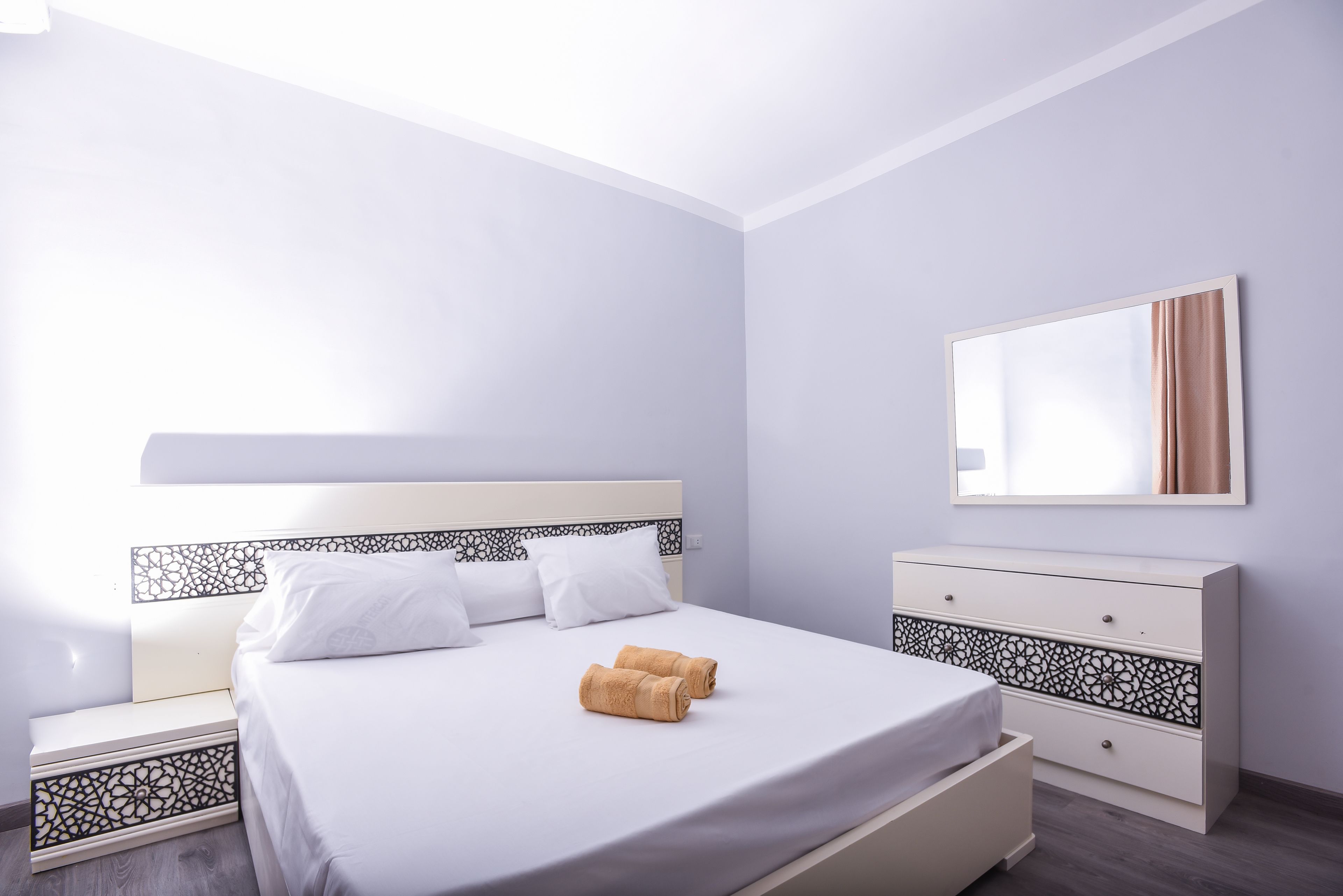 A neatly made bed with white linens and two rolled towels, a bedside table with a decorative panel, and a dresser with drawers.