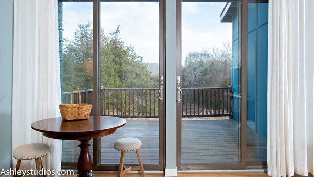 A room with a sliding glass door leading to a balcony, a round wooden table with a basket, and two cushioned stools. - Mid-Century Home Surrounded by Meadows (Pine Plains)