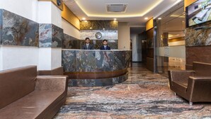 Un hall d’accueil d’hôtel avec un comptoir de réception en marbre, deux réceptionnistes et des sièges en cuir marron.