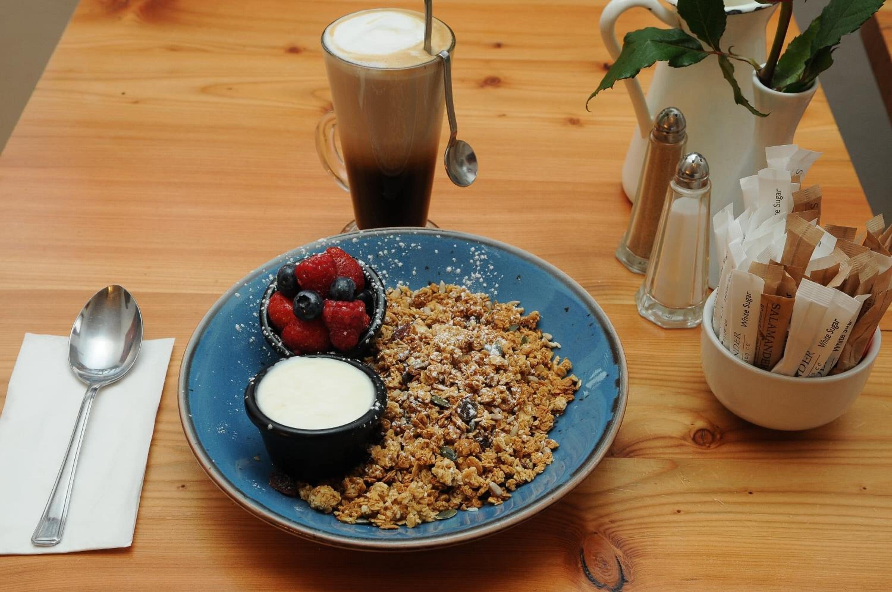 Une table en bois dressée pour le petit-déjeuner, avec une assiette de granola, des fruits rouges et du yaourt, une tasse de café et des sachets de sucre.