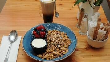 Une table en bois dressée pour le petit-déjeuner, avec une assiette de granola, des fruits rouges et du yaourt, une tasse de café et des sachets de sucre.