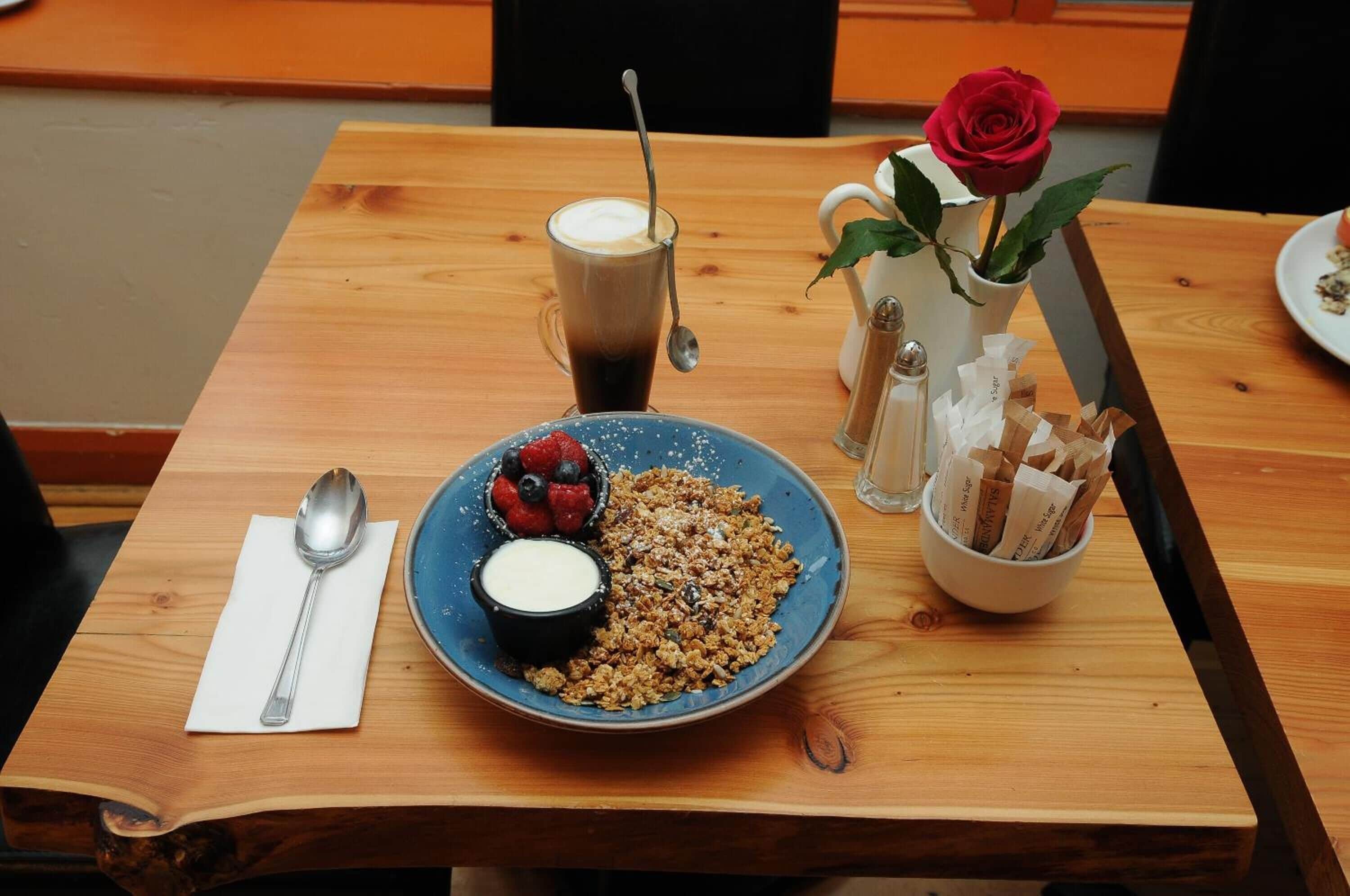 Une table en bois sur laquelle se trouvent une assiette de granola, une tasse de café, un bol de yaourt et un vase avec une rose rouge.