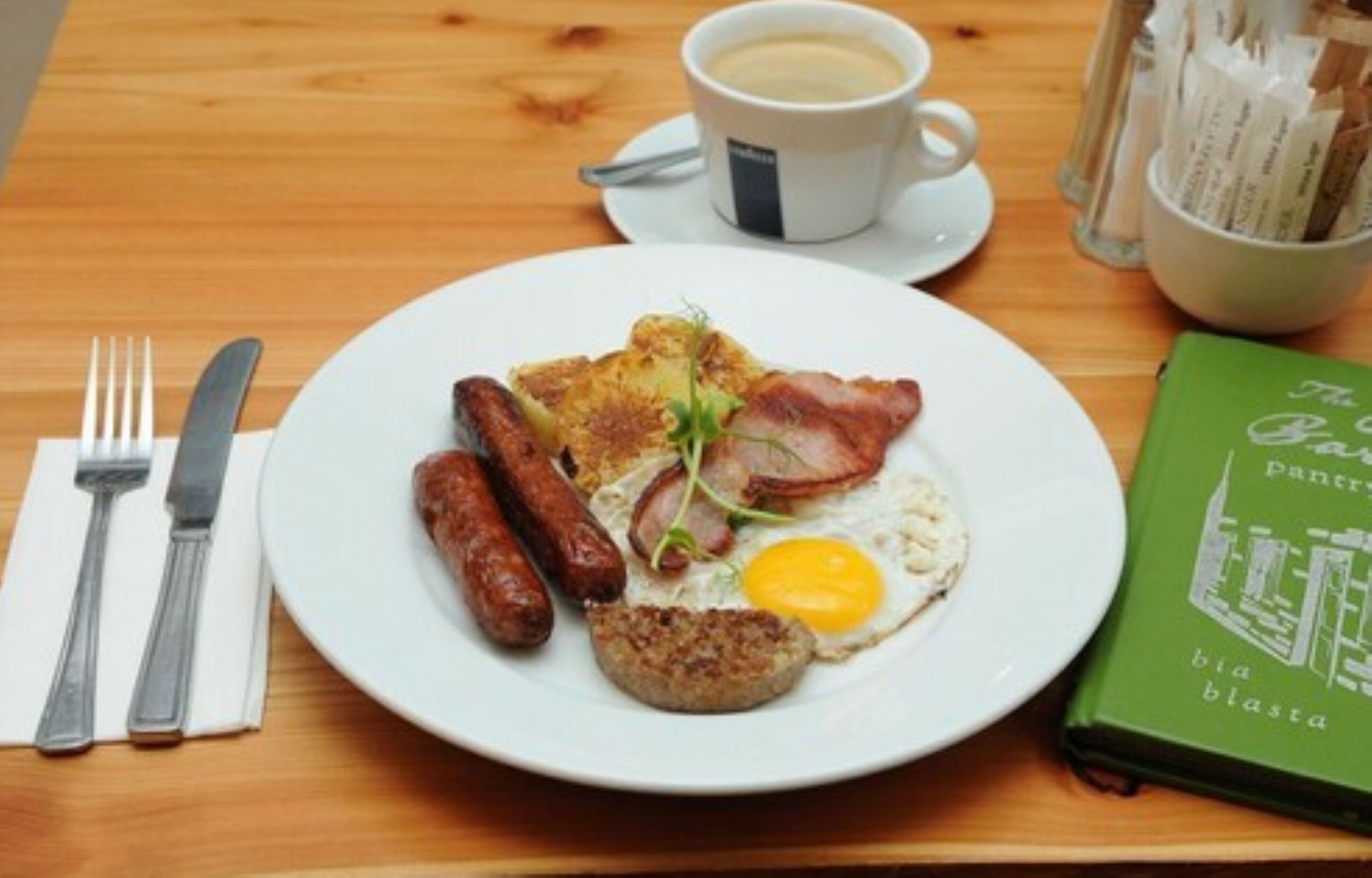 Une assiette de petit-déjeuner comprenant des saucisses, du bacon, un œuf au plat et des toasts, accompagnée d’une tasse de café et d’un livre intitulé « Le garde-manger du jardin ».