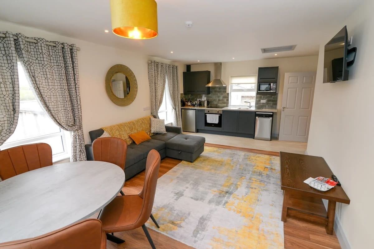 A modern living room with a grey sofa, a dining table with chairs, and a kitchen in the background.