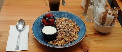 Un desayuno con un plato de granola, frutos rojos y yogur, una taza de café y condimentos sobre una mesa de madera.