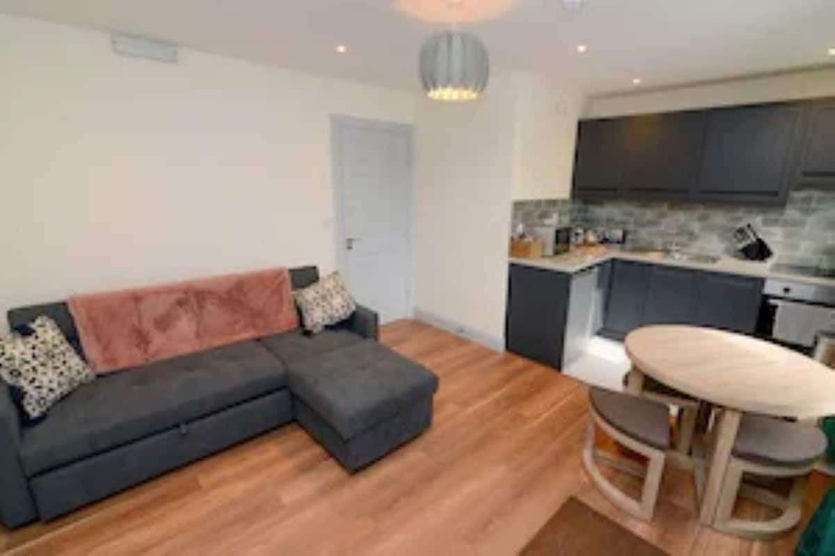 A modern living room with a grey sectional sofa, a small round table, and a kitchen area with dark cabinets and a stone backsplash.