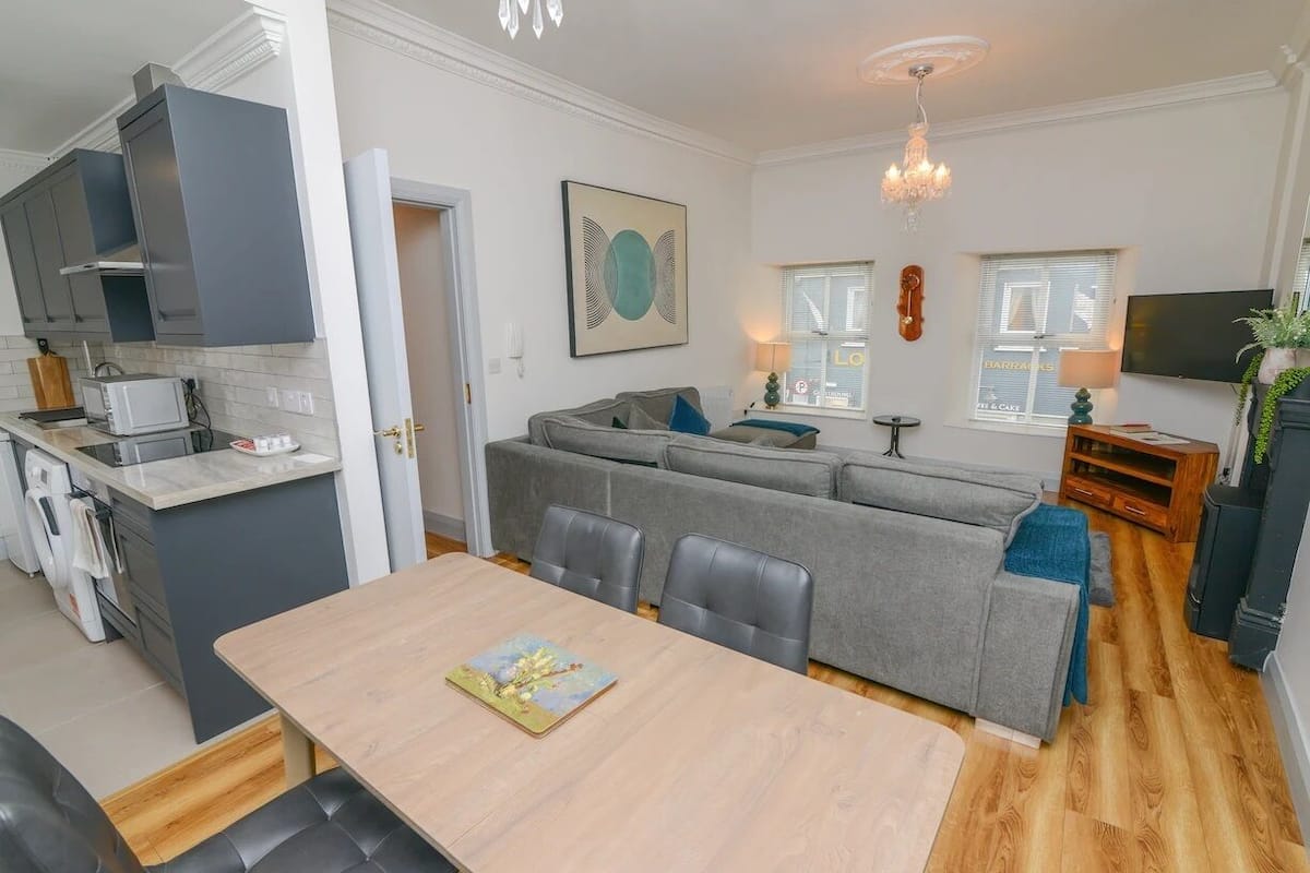 A modern kitchen with grey cabinets, a dining table with chairs, a grey sofa, and a chandelier.