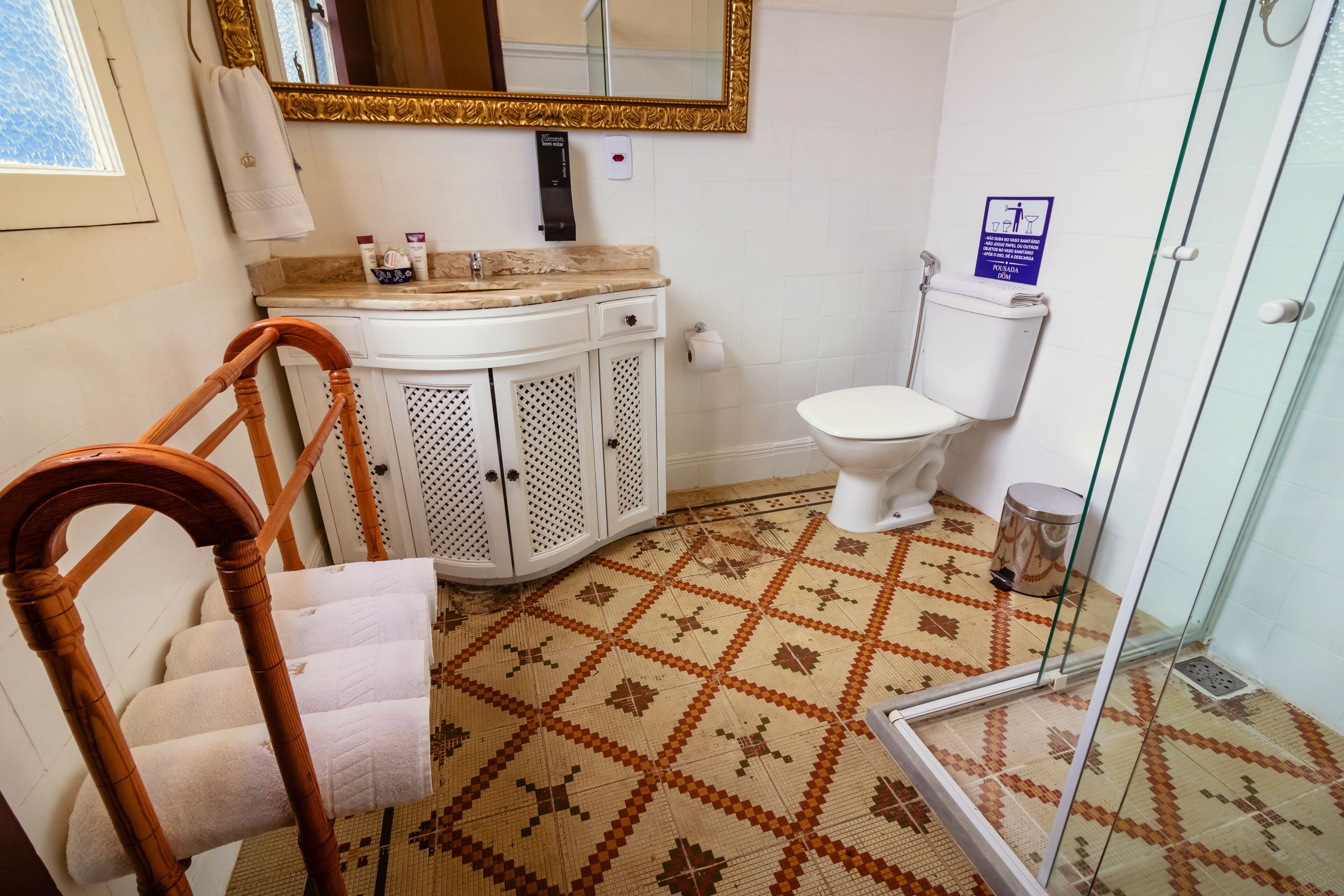 A bathroom with a patterned floor, a white toilet, a vanity with a mirror, and a wooden chair with towels.