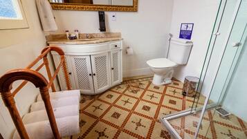 A bathroom with a patterned floor, a white toilet, a vanity with a mirror, and a wooden chair with towels.