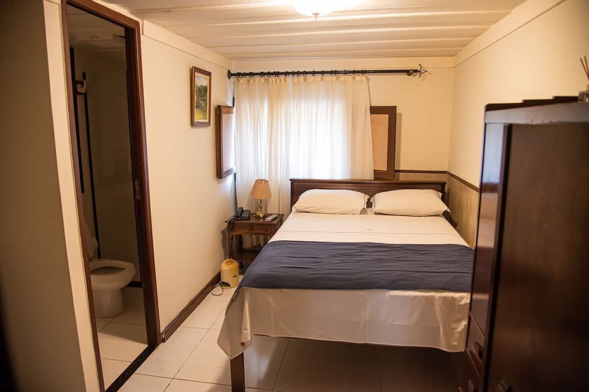 A bedroom with a bed, a bedside table with a lamp, a mirror, and a curtain.