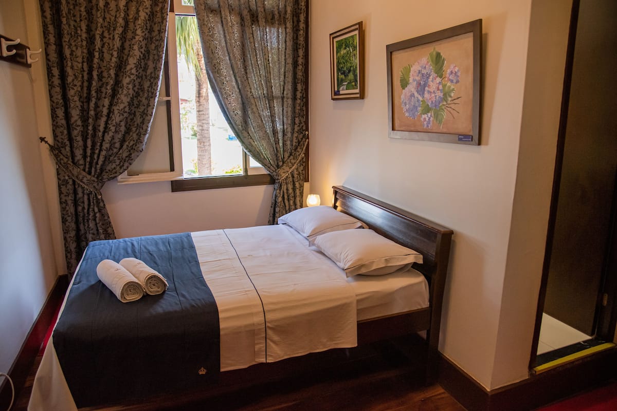 A neatly made bed with a blue bedspread and white linens, a window with curtains, and two framed floral paintings on the wall.