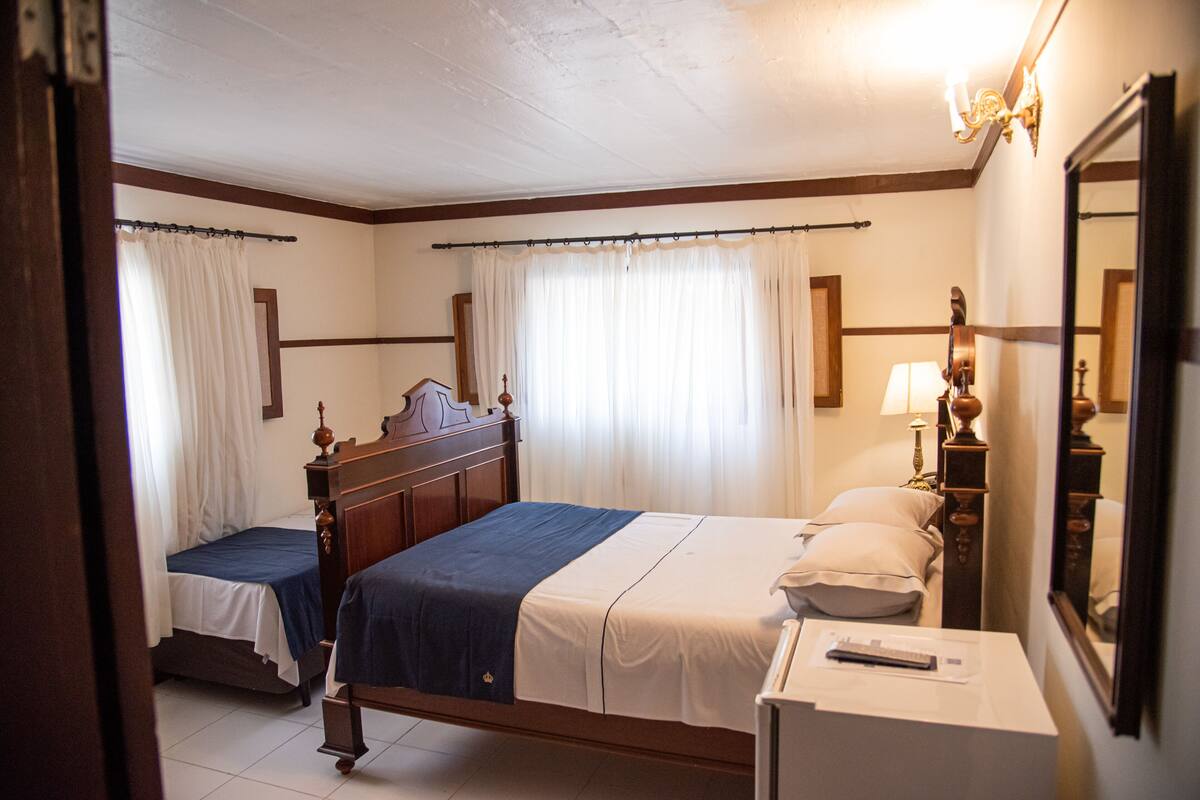 A hotel room with two beds, a bedside table with a lamp, a mirror, and a window with curtains.